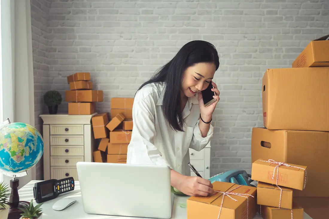 An online seller on the phone, labelling parcels among the boxes in her stockroom.